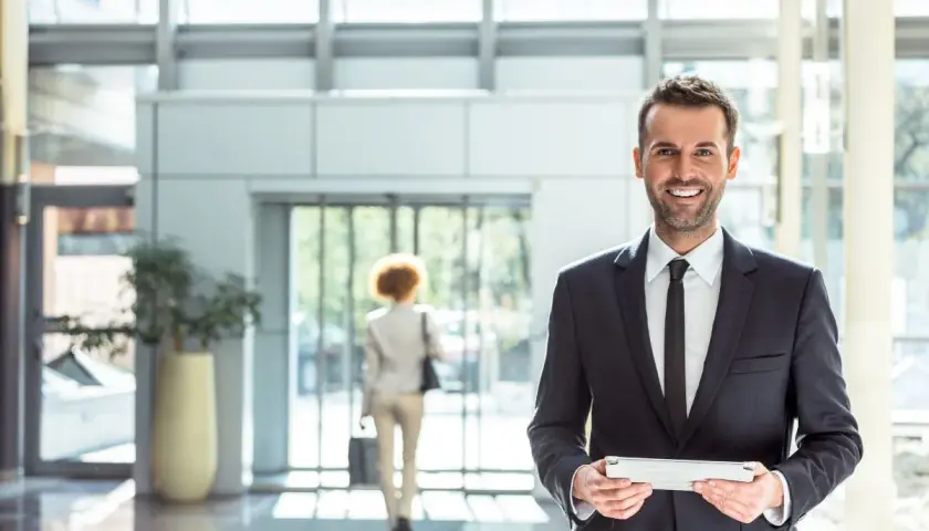 Uomo sorridente in abito scuro con tablet, in un moderno atrio di un pms hotel.