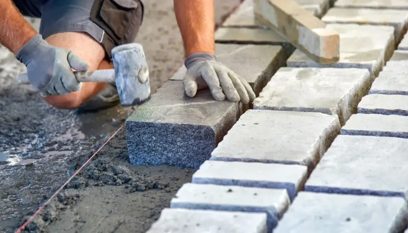 Układanie kostki brukowej na podsypce. Ręce w rękawiczkach kładą kamień, używając gumowego młotka. Widać suchy beton proporcje.
