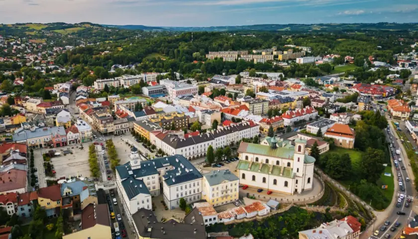 Urokliwe miasto w dolinie ropy, z dominującą białą bazyliką i kolorowymi kamienicami, otoczone zielonymi wzgórzami.