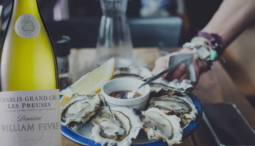 Une bouteille de Chablis Grand Cru William Fèvre, parfaite pour savoir quoi boire avec des huîtres. Une assiette d'huîtres fraîches et un quartier de citron complètent la scène.