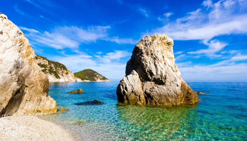 Plage de galets et rochers imposants dans une eau turquoise cristalline, évoquant une charmante ville toscane bord de mer sous un ciel bleu éclatant.