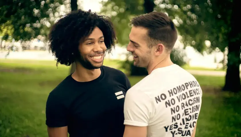 Due uomini sorridono, uno con maglia nera con simbolo di uguaglianza, l'altro con maglia bianca con messaggi di inclusione. Un'immagine che evoca il poliamore significato di amore libero e rispetto.