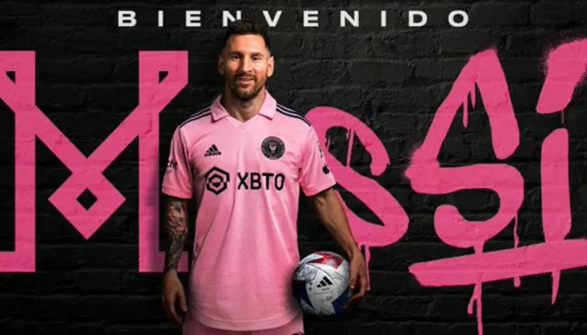 Lionel Messi w różowej koszulce Inter Miami, z piłką w ręku, na tle napisu "Bienvenido Messi". Nowy inter miami trener.