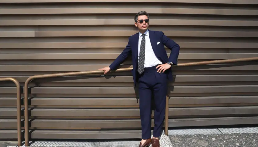 Un homme élégant porte un costume bleu marine, une chemise blanche et une cravate à motifs. Ses chaussures marron complètent parfaitement sa tenue.