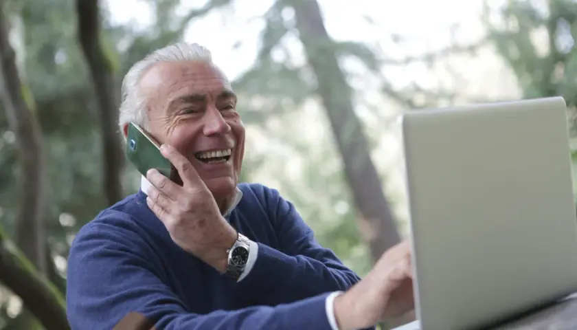 Uśmiechnięty starszy mężczyzna rozmawia przez telefon i pracuje na laptopie na zewnątrz.