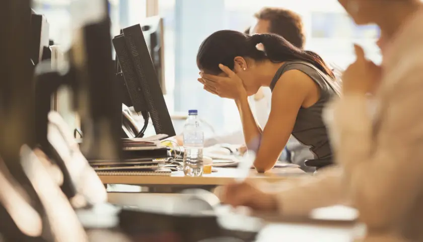 A woman sits at her desk, head in hands, looking demoralized at work. A bottle of water sits nearby.