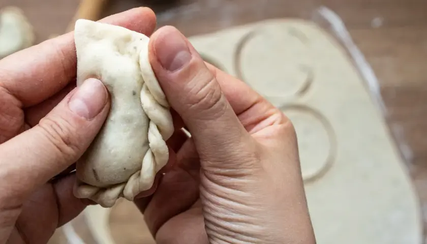 Dłonie formują brzeg pieroga, pokazując, jak lepić pierogi. W tle rozwałkowane ciasto z wyciętymi kółkami.