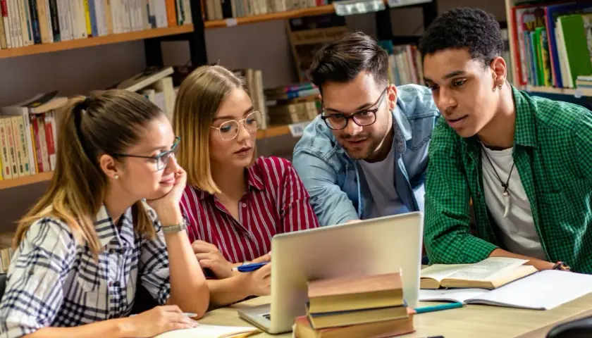 Studenci na studiach humanistycznych pochylają się nad laptopem w bibliotece, dyskutując o projekcie.