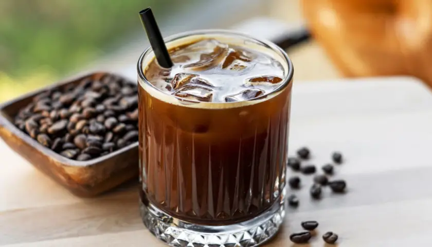 Erfrischender kalter Kaffee mit Eiswürfeln und einem Strohhalm, daneben eine Schale mit Kaffeebohnen.