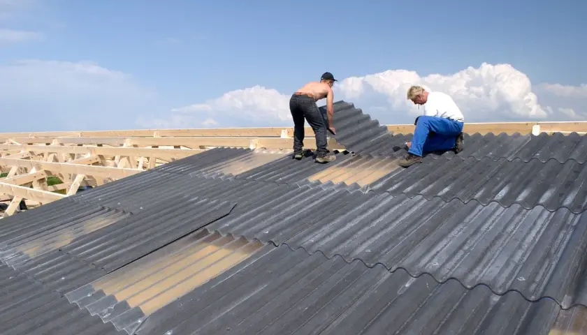 Zwei Männer verlegen graue Faserzementplatten auf einem Dach. Die Wellplatten werden auf einer Holzkonstruktion befestigt.