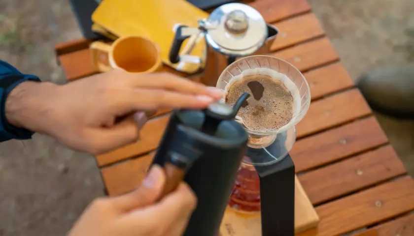 Ręce przygotowują aromatyczną kawę w górach. Na drewnianym stoliku stoi dripper z kawą, obok czajniczek i kubek.