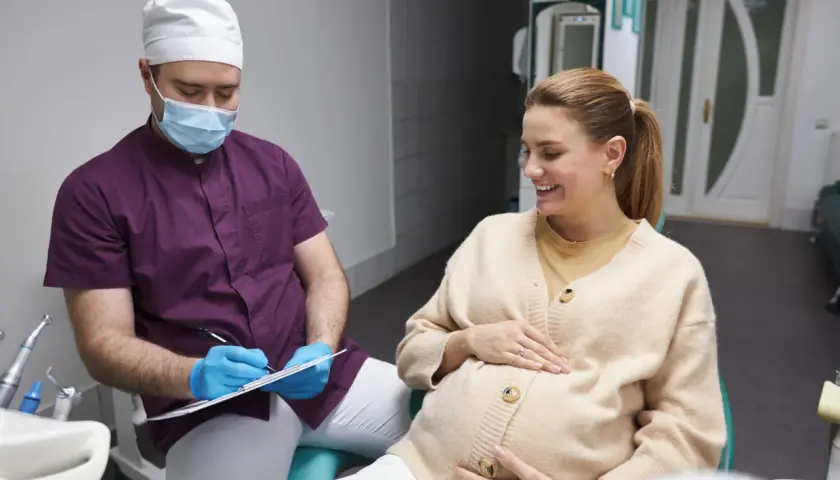 Dentista con mascherina parla con una donna incinta sorridente, discutendo della pulizia dei denti in gravidanza.