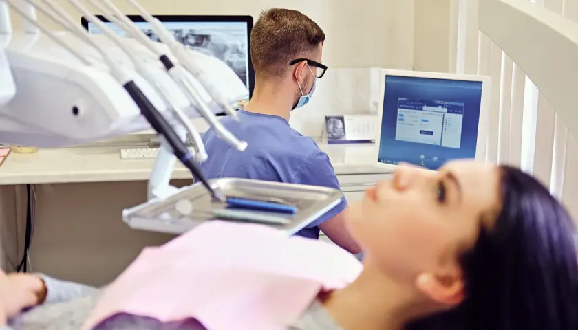 Un dentista con mascherina e occhiali lavora in uno studio dentistico moderno, esaminando radiografie su un monitor.