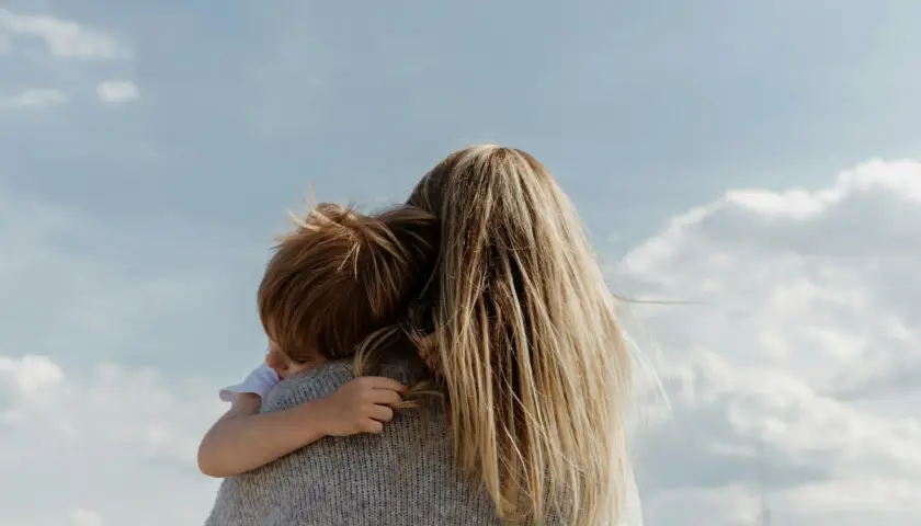 En mamma kramar sitt barn under en blå himmel med moln. En varm kram mellan mammor och minimodeller.
