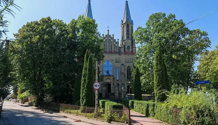 Neogotycki kościół w Krynkach, otoczony zielenią, z wieżami i charakterystycznym oknem.