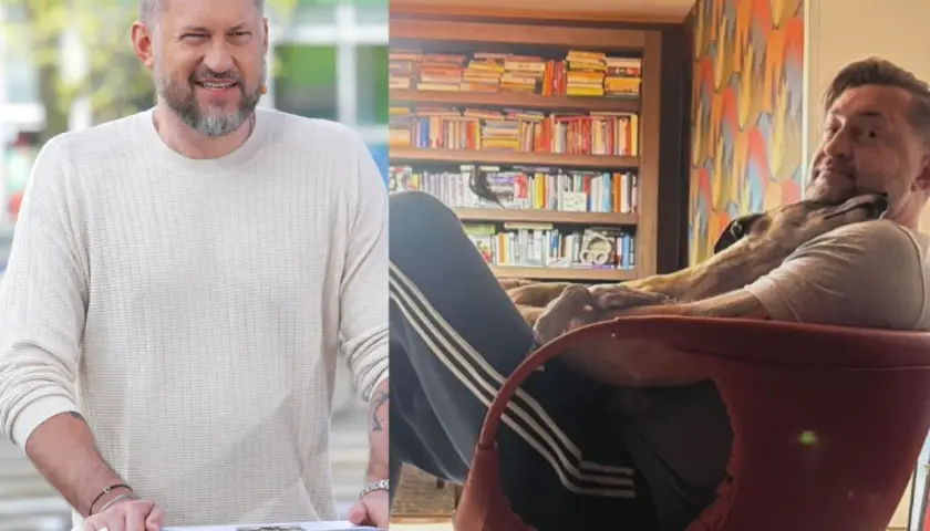 Marcin Prokop w swetrze i z psem na kolanach na tle regału z książkami.