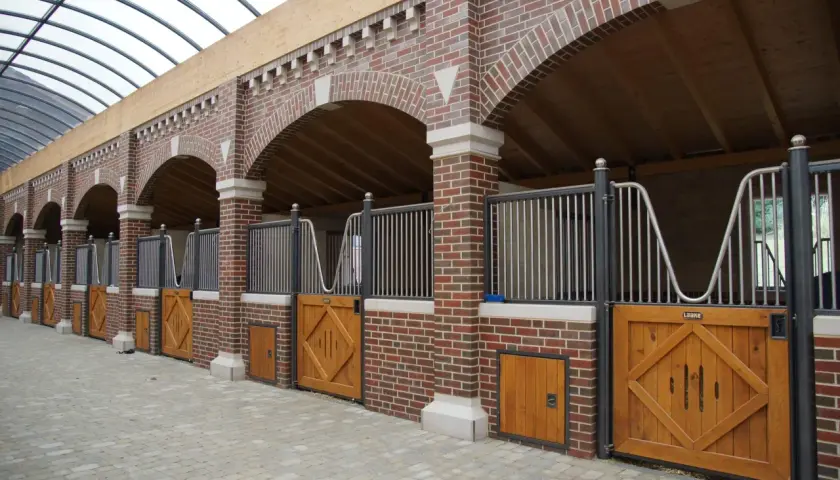 Rangée de stalles de chevaux en briques avec portes en bois et toiture transparente.