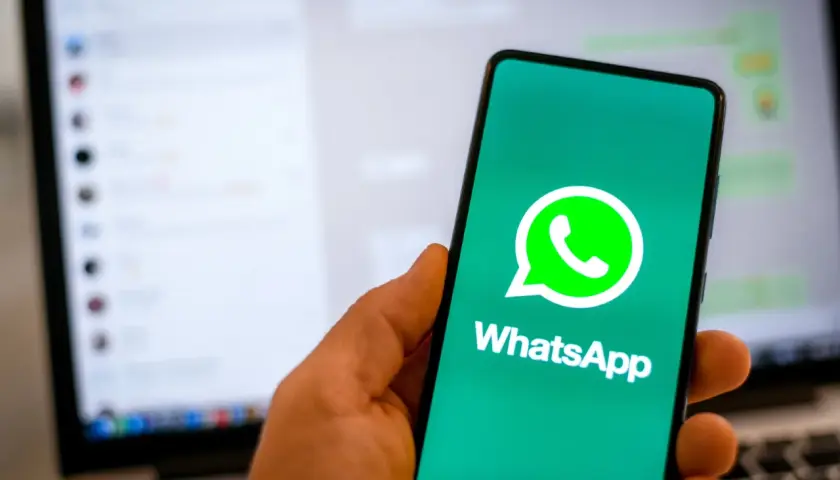 Dłoń trzyma smartfon z logo WhatsApp. W tle widać ekran laptopa z otwartym programem do komunikacji, sugerującym użycie WhatsApp na komputer.