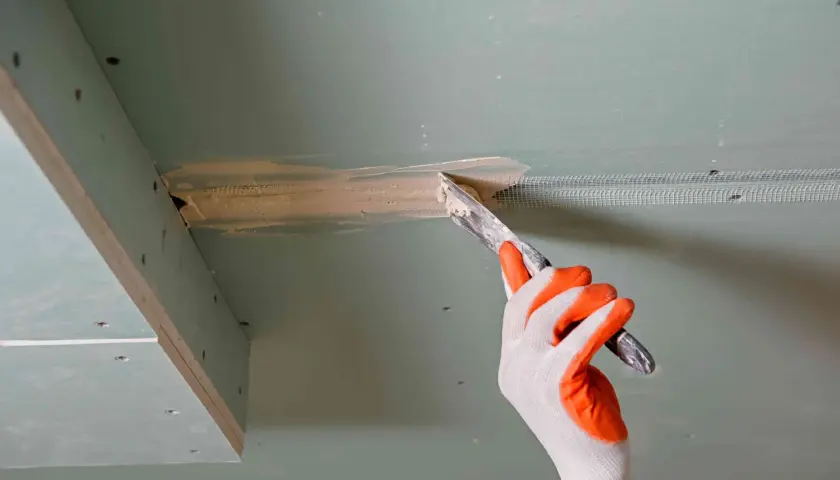 Ręka w rękawicy nakłada masę szpachlową na połączenie płyt gipsowo-kartonowych, przygotowując je do dalszych prac.
