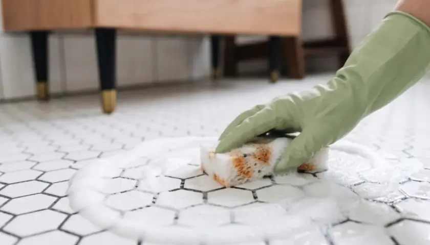 Rękawiczka w zielonej gumie czyści pianą białe, matowe płytki podłogowe.