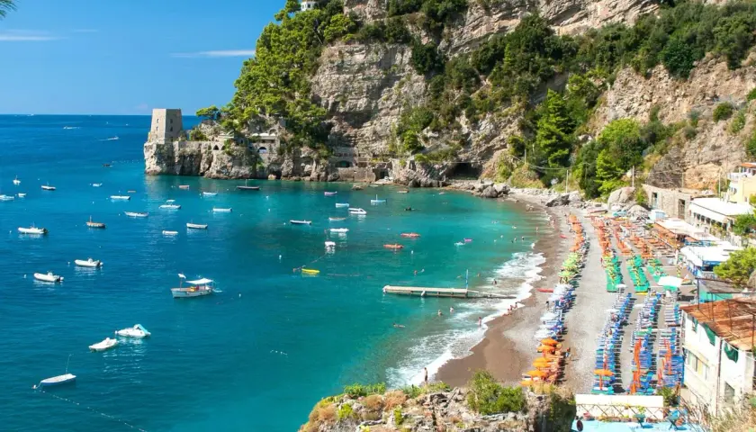 Vue panoramique d'une plage de galets animée à Almafi, Italie, avec des parasols colorés, des chaises longues et des eaux bleues claires parsemées de bateaux.