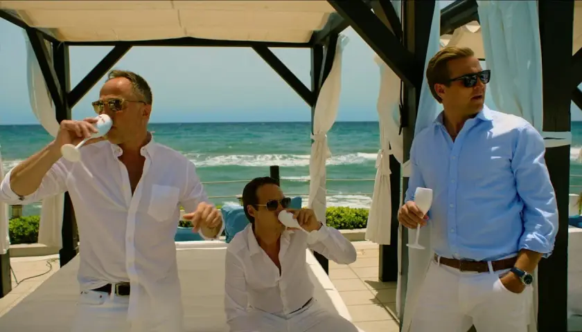 Tre män i vita och ljusblå skjortor dricker ur champagneglas vid havet. En scen från en exit serie.