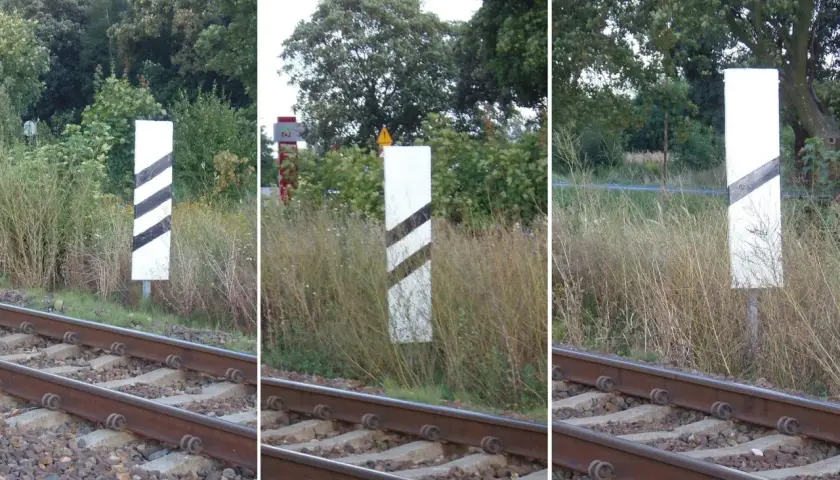 Trzy białe słupki z czarnymi pasami, wskaźnik w11a, stoją przy torach kolejowych.