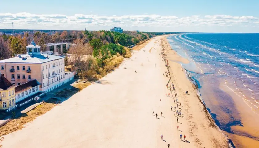 Szeroka, piaszczysta plaża na Łotwie, z budynkami przy brzegu i ludźmi spacerującymi wzdłuż morza.