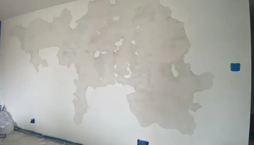 Odpada farba ze ściany, odsłaniając tynk. Na podłodze resztki farby i niebieska taśma.