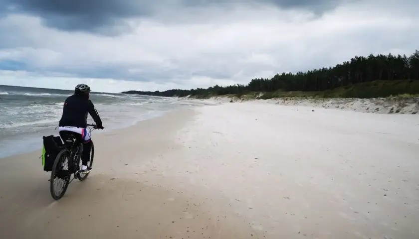 Rowerzystka przemierza piaszczystą plażę na mierzei sarbskiej, z widokiem na wzburzone morze i sosnowy las.