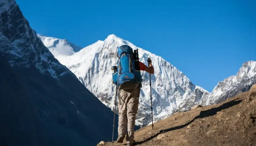 Un excursionista con mochila azul asciende un sendero polvoriento, bastones en mano, ante majestuosas montañas nevadas. Una aventura de trekking Manaslu.