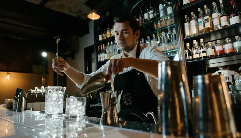Barman przygotowuje drinka, zastanawiając się, ile zarabia dobry barman. Za nim półki z alkoholem.