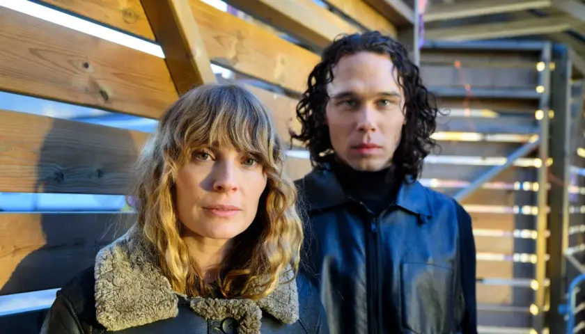 Två personer, en kvinna med blont hår och en man med lockigt mörkt hår, står framför en träkonstruktion. De är skådespelare från rollistan i Knutby (tv-serie).