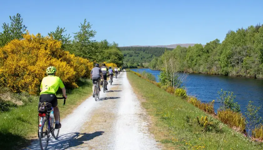 A group enjoys cycling in Scotland along a canal path, with bright yellow gorse bushes and lush green trees lining the route.