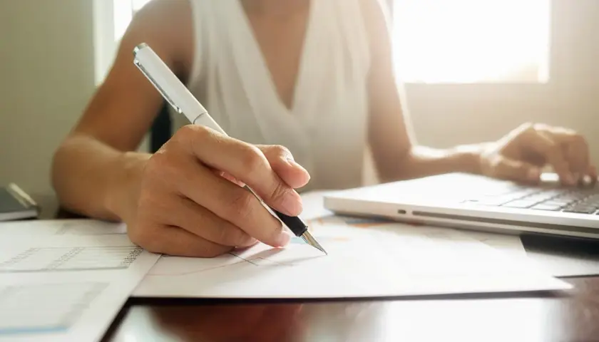 Osoba pisze długopisem na papierze, obok laptopa. Tworzy regulamin grupy na FB, dbając o szczegóły.