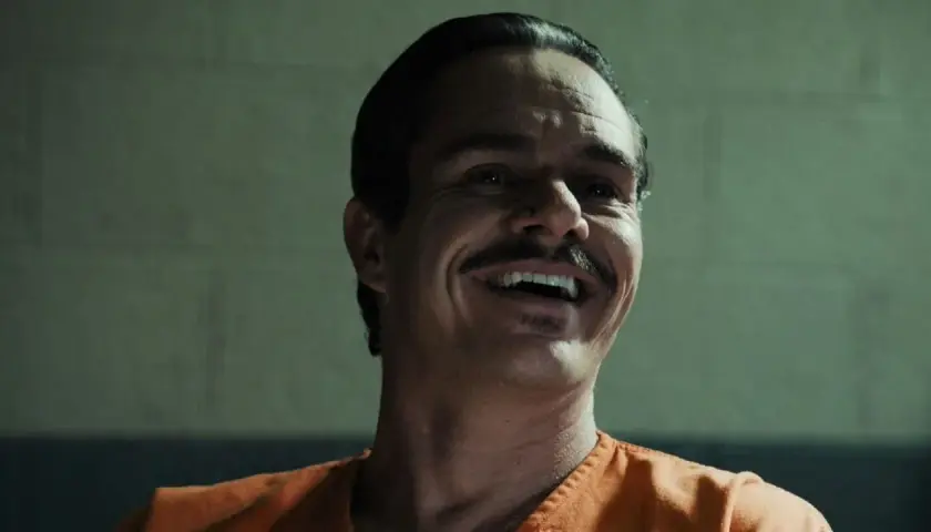 Lalo Salamanca ler skrattande i en orange fängelsedräkt, hans mustasch framträder tydligt.