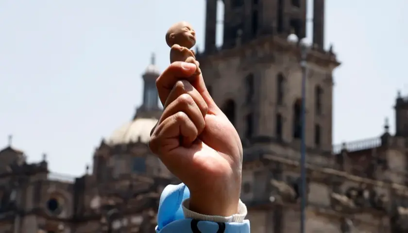 Hand hält Fötus-Figur vor Kirche. Debatte um evangelische Kirche und Abtreibung.
