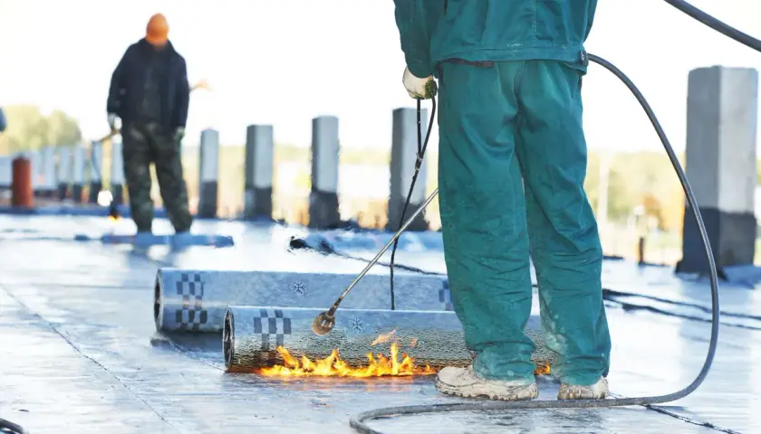Arbeiter schweißen Bitumenbahnen, um ein Flachdach abzudichten. Flammen erhitzen das Material für eine sichere Verbindung.