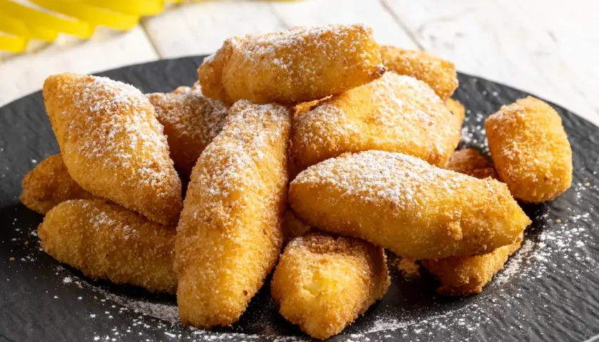 Un mucchio di dorate crema fritta, cosparse di zucchero a velo, su un piatto scuro.