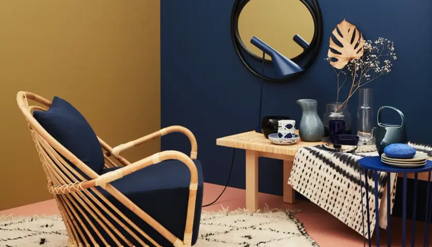 Ein Rattan-Sessel mit dunkelblauem Kissen steht auf einem flauschigen Teppich. Welche Farbe passt zu Gold? Hier: ein Spiegel mit goldenem Rahmen und ein gelber Wandbereich.