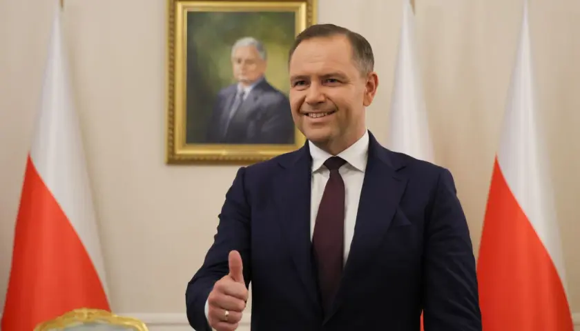 Mężczyzna w garniturze pokazuje kciuk w górę na tle portretu i flag Polski.