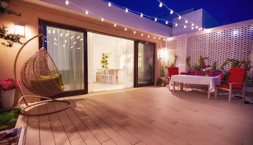 Terraza con silla colgante y mesa, iluminada por una guirnalda de luces. La pantalla lámpara plisada ilumina el ambiente.