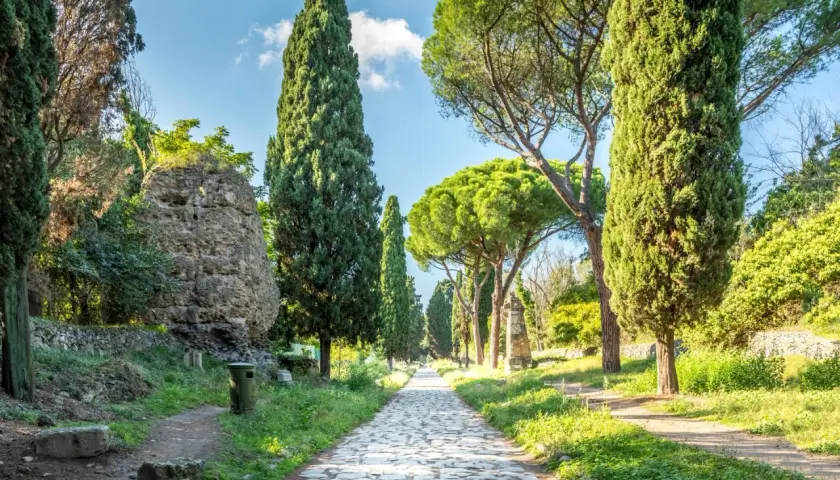 Allée pavée bordée de cyprès et de pins sur la Via Appia, avec des ruines antiques sur le côté.