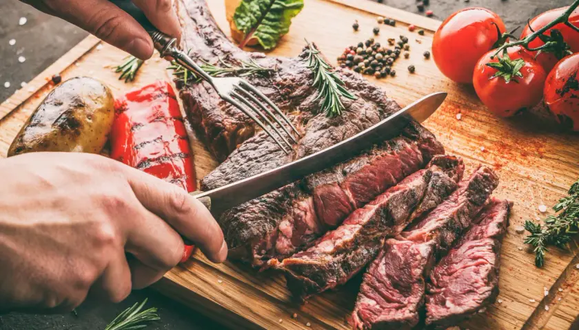 Une main coupe un succulent côte de bœuf barbecue sur une planche en bois, accompagné de pommes de terre rôties et de poivrons grillés.