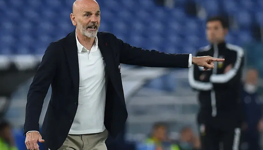 Trener Stefano Pioli w garniturze i białej koszulce polo wskazuje palcem, udzielając wskazówek na boisku.