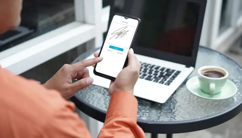 Persona usando su móvil para firmar online documento. En la mesa hay un portátil y una taza de café.