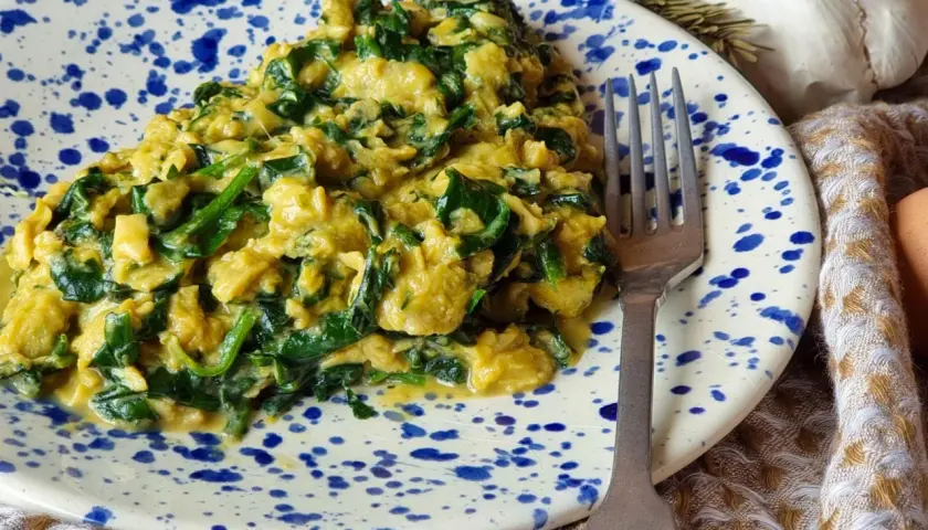 Delicioso revuelto de espinacas cremoso y sabroso, servido en un plato con salpicaduras azules. Un tenedor plateado descansa a su lado.