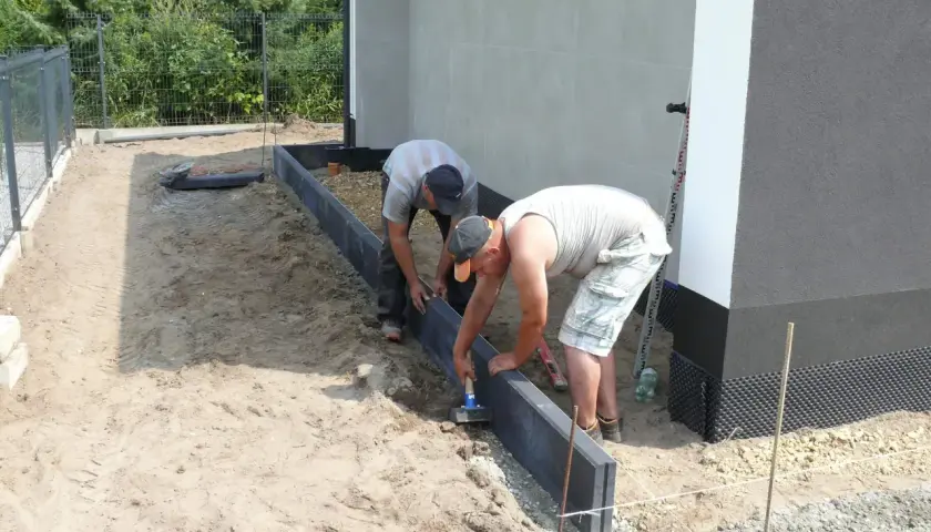 Dwóch mężczyzn montuje obrzeża. Używają młotka do osadzenia ich w suchym betonie, przygotowując teren pod przyszły ogród.