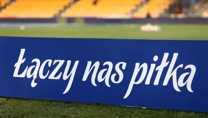 Niebieska tablica z napisem "łączy nas piłka" na tle stadionu. To motto, które jednoczy kibiców i prezesa PZPN.