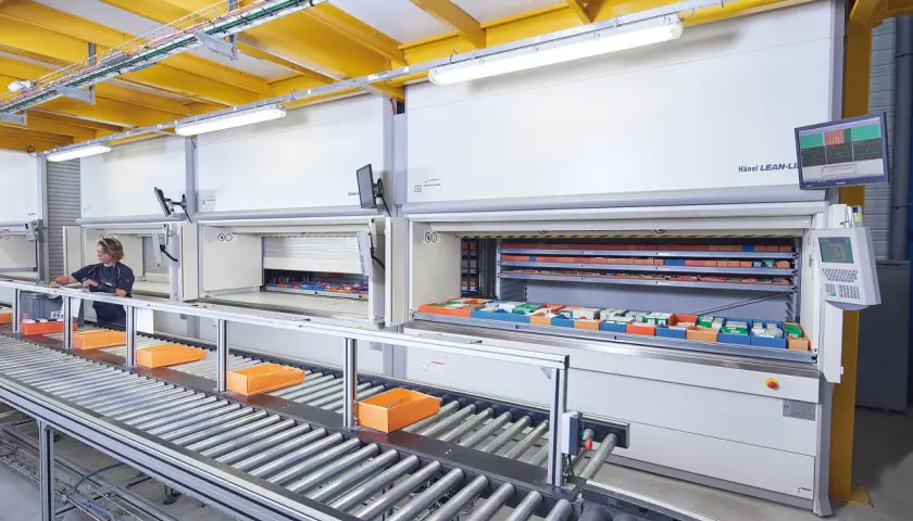 Ein Kommissionierlager mit automatisierten Lagersystemen. Eine Mitarbeiterin entnimmt Teile, während Behälter auf einem Rollenband transportiert werden.